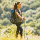Genouillère Orliman Flexilig Max pour traumatismes du genou, portée par une jeune femme souriante en randonnée avec sac à dos et bâton de marche, dans un paysage naturel ensoleillé
