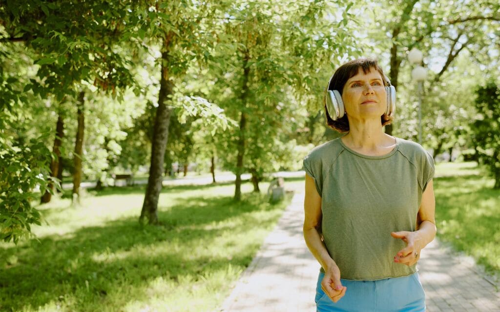 Femme d'âge mûr pratiquant la marche active dans un parc avec des écouteurs pour prévenir les douleurs articulaires.