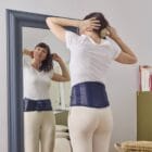 Femme debout se regardant dans le miroir de sa chambre avec une ceinture lombaire de thermothérapie Prim’Arthrose pour soulager les douleurs liées à l’arthrose lombaire débutante