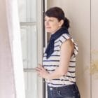 Femme debout près d'une fenêtre avec autour du cou, l’écharpe de thermothérapie Prim’Arthrose pour soulager les douleurs liées à l’arthrose cervicale débutante