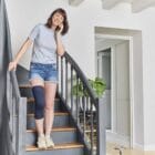 Femme au téléphone, debout dans ses escaliers et portant la genouillère rotulienne de confort Prim’Arthrose pour soulager les douleurs liées à la gonarthrose débutante